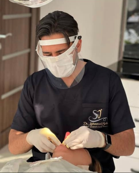 A male dentist wearing a black scrub top with a logo on the chest pocket and a face mask, performing a dental surgery.