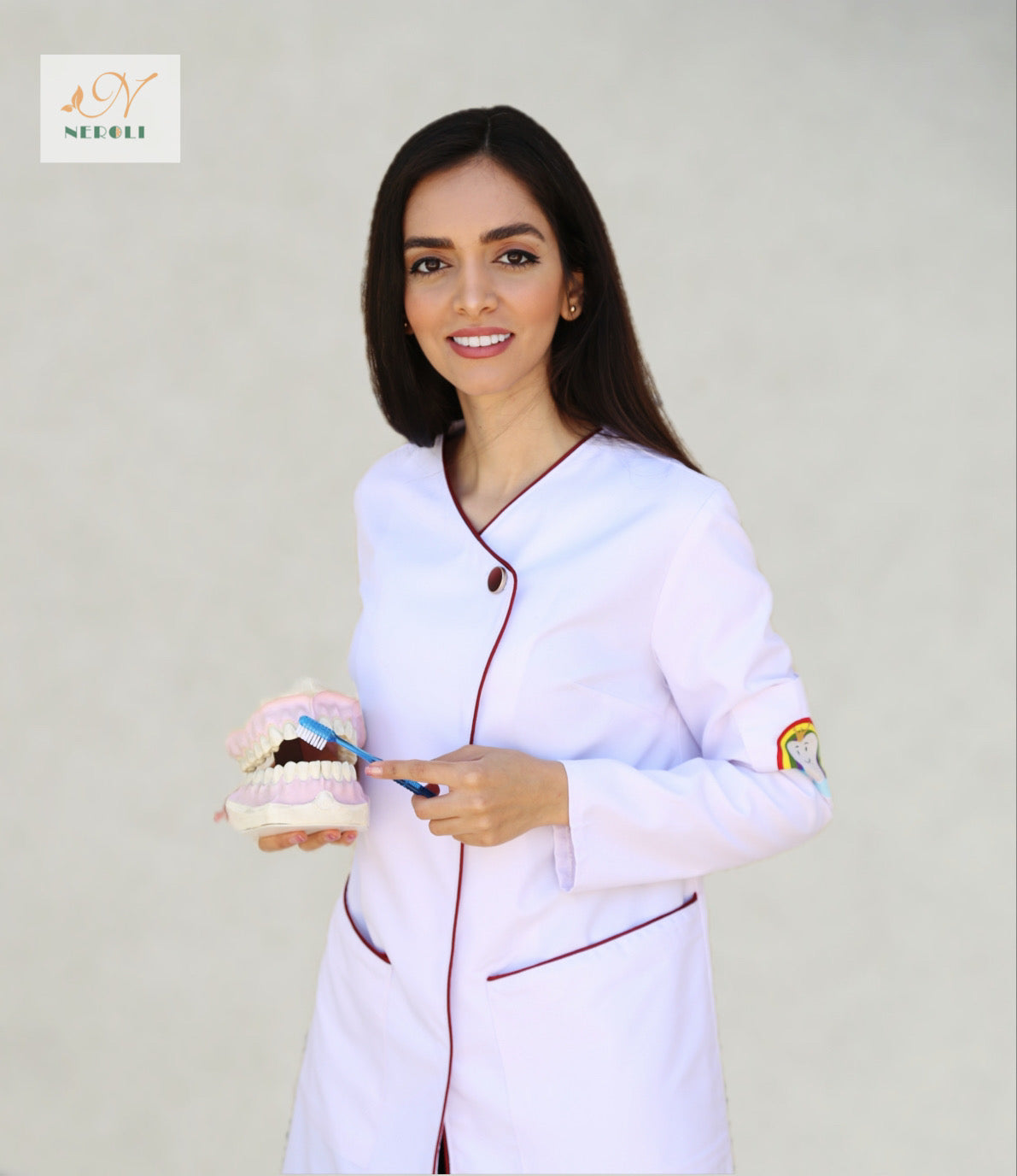 A female dentist in lab coat with dark red piping, holding a toothbrush and duplicate teeth set.